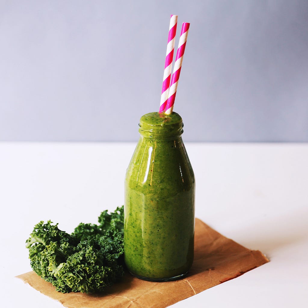 Green smoothie in a clear bottle with 2 pink and white straw, sitting next to kale.