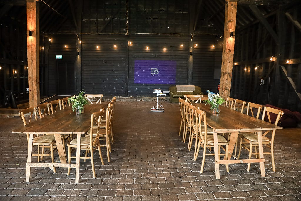 The main barn at Elmley with two sets of long trestle tables with chairs and flowers on them with the Black design Guild logo on a purple background projected on to the barn wall.