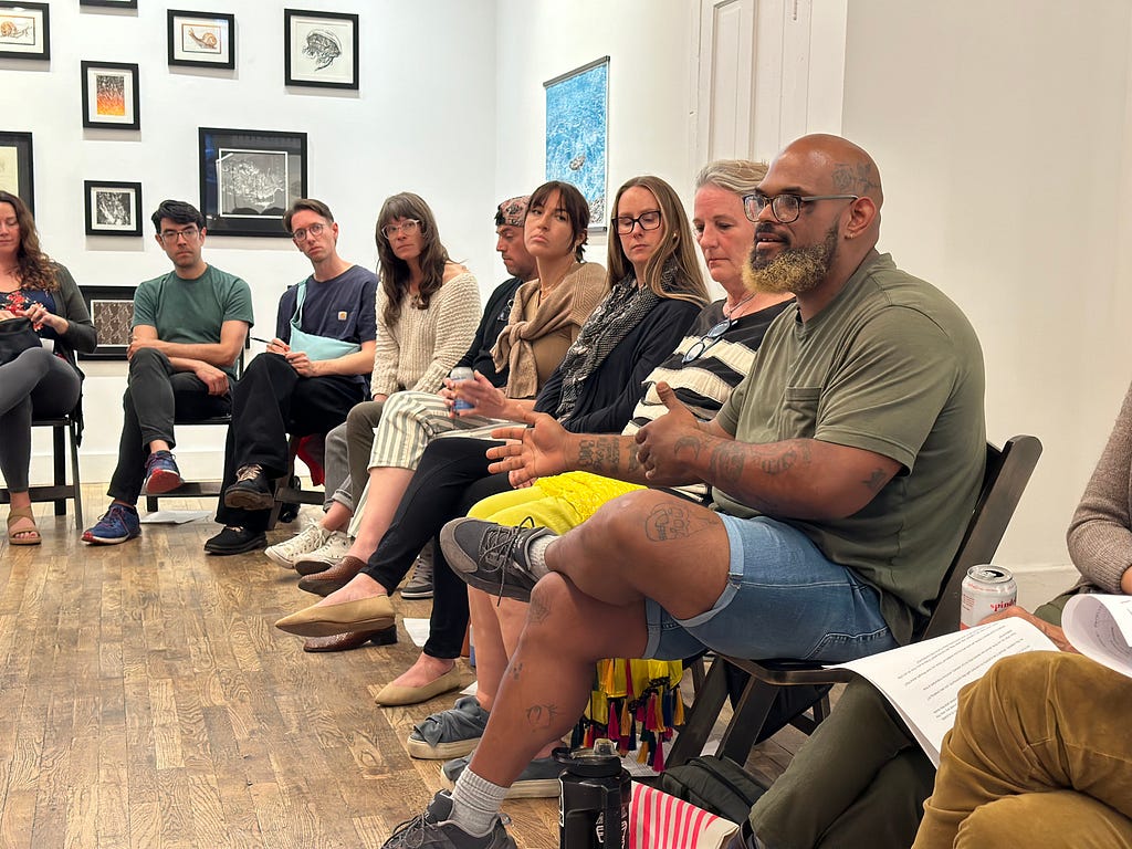 Nine people seated in a semi-circle are visible but part of a larger group. In the front of the frame an African-American man with glasses and a grey beard gesticulates with his arms as he speaks to the group. He wears jean shorts and an army green t-shirt. Others listen. Framed artworks hang on the walls behind the group.