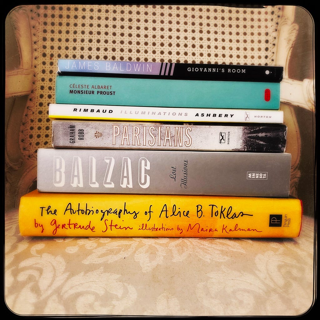 A stack of books about Paris include Giovanni’s Room by James Baldwin, Monsieur Proust by Céleste Albaret, Illuminations by Arthur Rimbaud, Parisians by Graham Robb, Lost Illusions by Honoré de Balzac, and The Autobiography of Alice B. Toklas by Gertrude Stein.