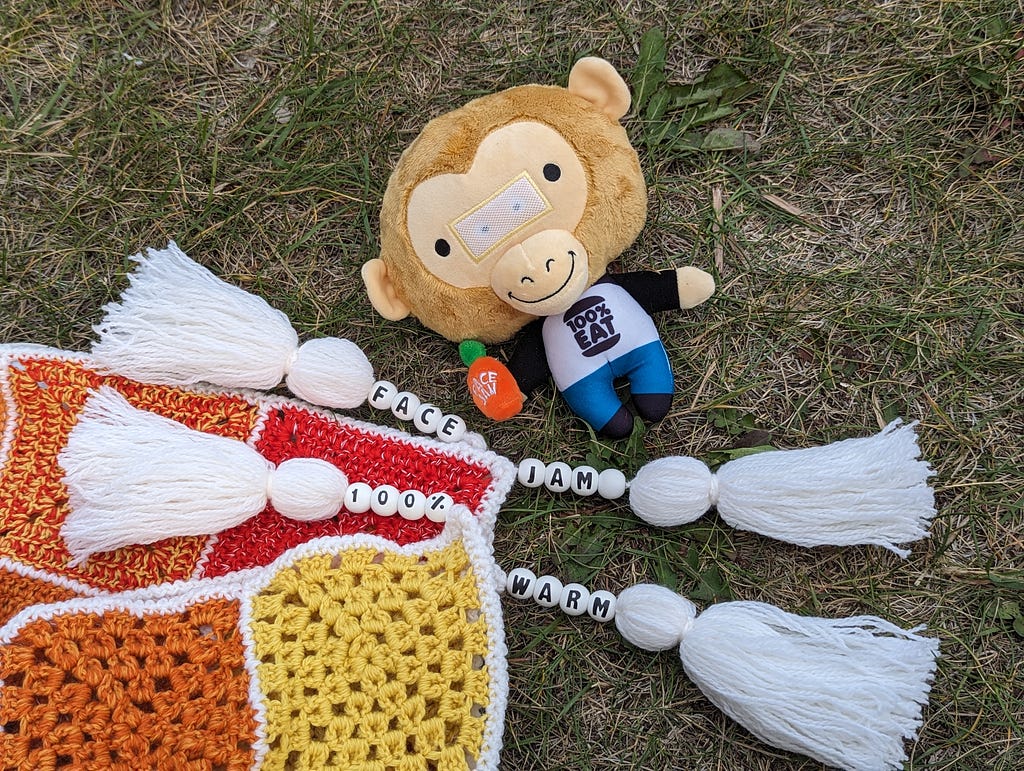 Sauce Monkey plush lays just above the four tassels of the blanket. Letter beads on the tassels spell out FACE JAM 100% WARM.
