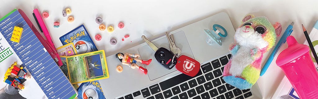 A laptop covered in bits and pieces of parenthood and school, including a car key with a university keychain, cereal, action figures, and a pacifier.