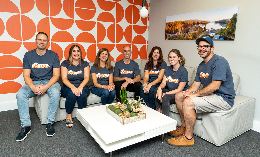 Seven members of the Architectural Designs team pose in an office with a bright orange and write wall. They are wearing blue shirts that read Bueno to honor AD alumnus Ryan Bard, who is pursuing his lifelong dream of becoming a commercial pilot.