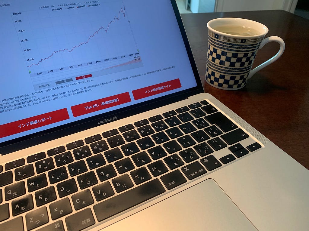 A laptop displaying a financial chart with a steady upward trend on the screen. The keyboard of the laptop is in focus, and a blue and white patterned coffee mug is placed beside it on the table. The screen shows buttons labeled “India Related Reports,” “The BiC (News and Information),” and “India Investment Sites.