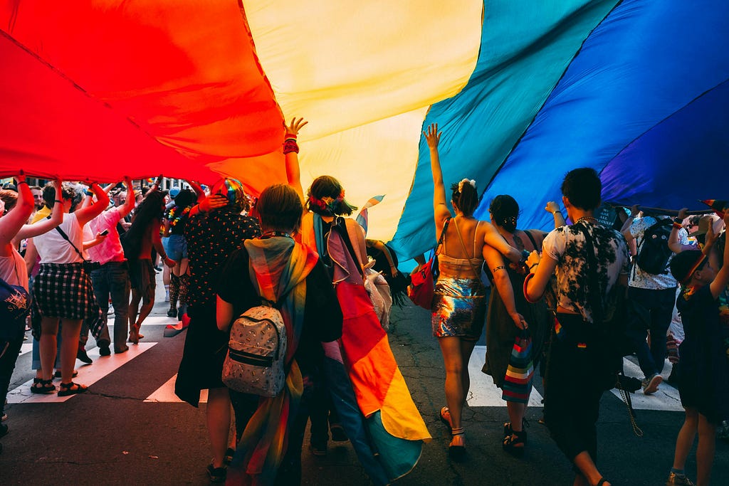 LGBT+ Supporters marching under a big rainbow LGBT Flag