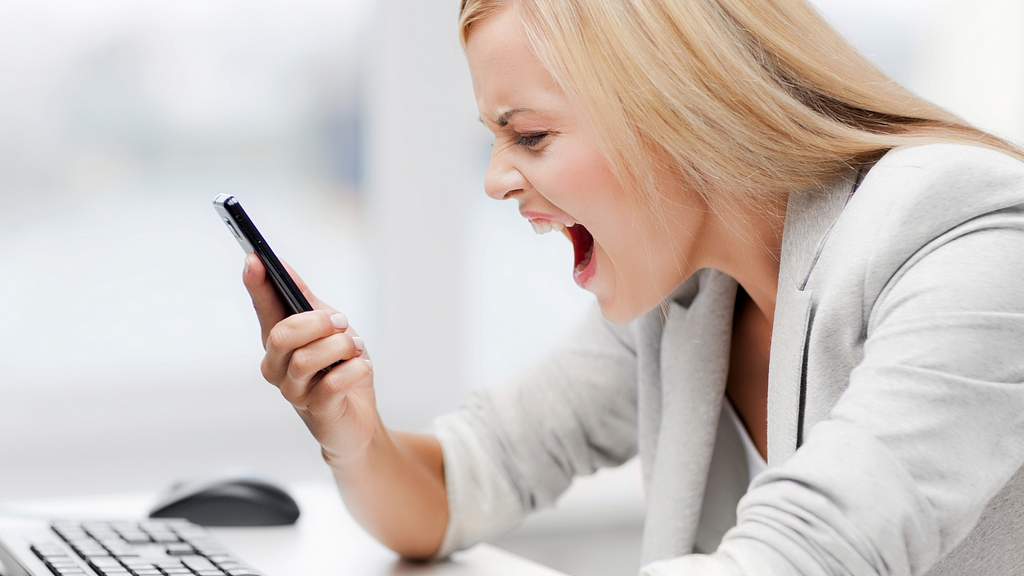 Frustrated blonde woman in a grey suit jacket yelling at her cell phone.