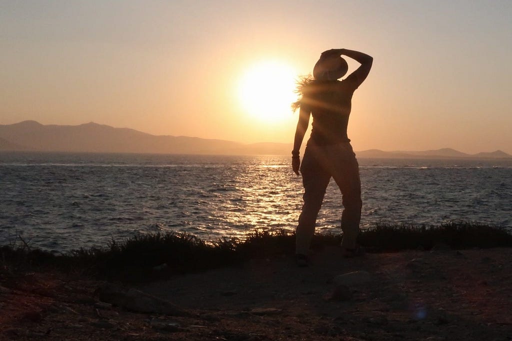 A sun sets over mountains on a horizon behind water. In the forefront is the silhouette of a woman standing on grass, gazing out over the water, with one arm raised holding on to her hat.