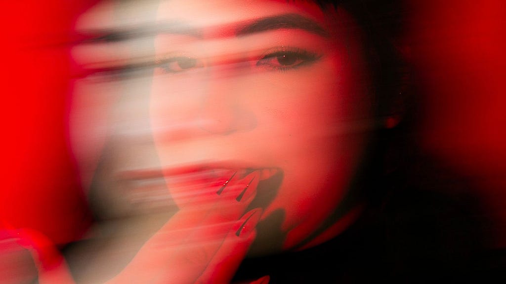 A blurred, artistic portrait of a person with dark hair, makeup, and long nails against a vivid red background. The motion blur creates a dynamic, ghostly effect, making the face and fingers appear multiple times in a horizontal direction.