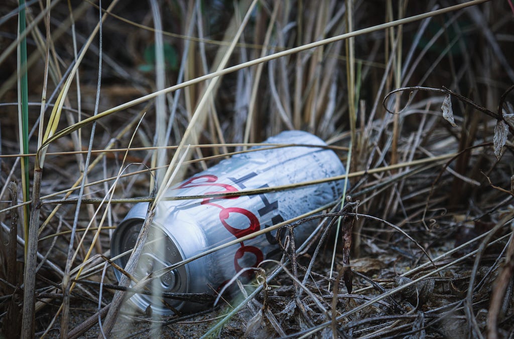 A discarded silver can of Coors Light beer lies on the ground amidst weeds and dirt.