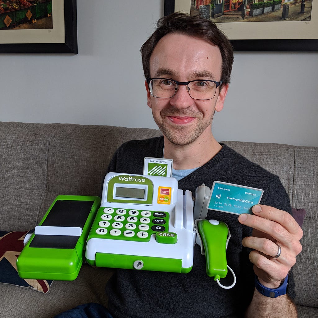 A smiling man (the author) holding up a Waitrose toy cash register and a John Lewis / Waitrose Partnership Card