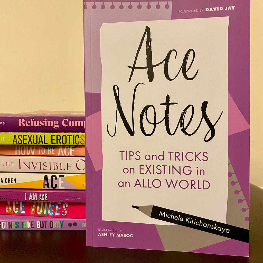 A book (“Ace Notes: Tips and Tricks on Existing in an Allo World”) placed in front of a stack of ace-themed books.