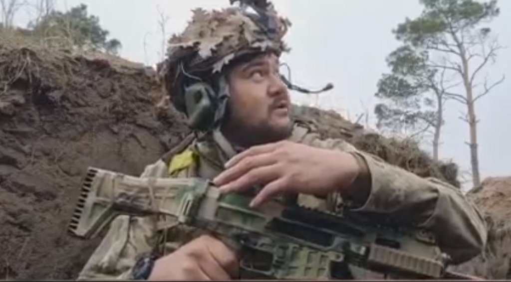 Ukrainian soldier in full military gear, looks up toward sky, from a ditch or place of hiding.