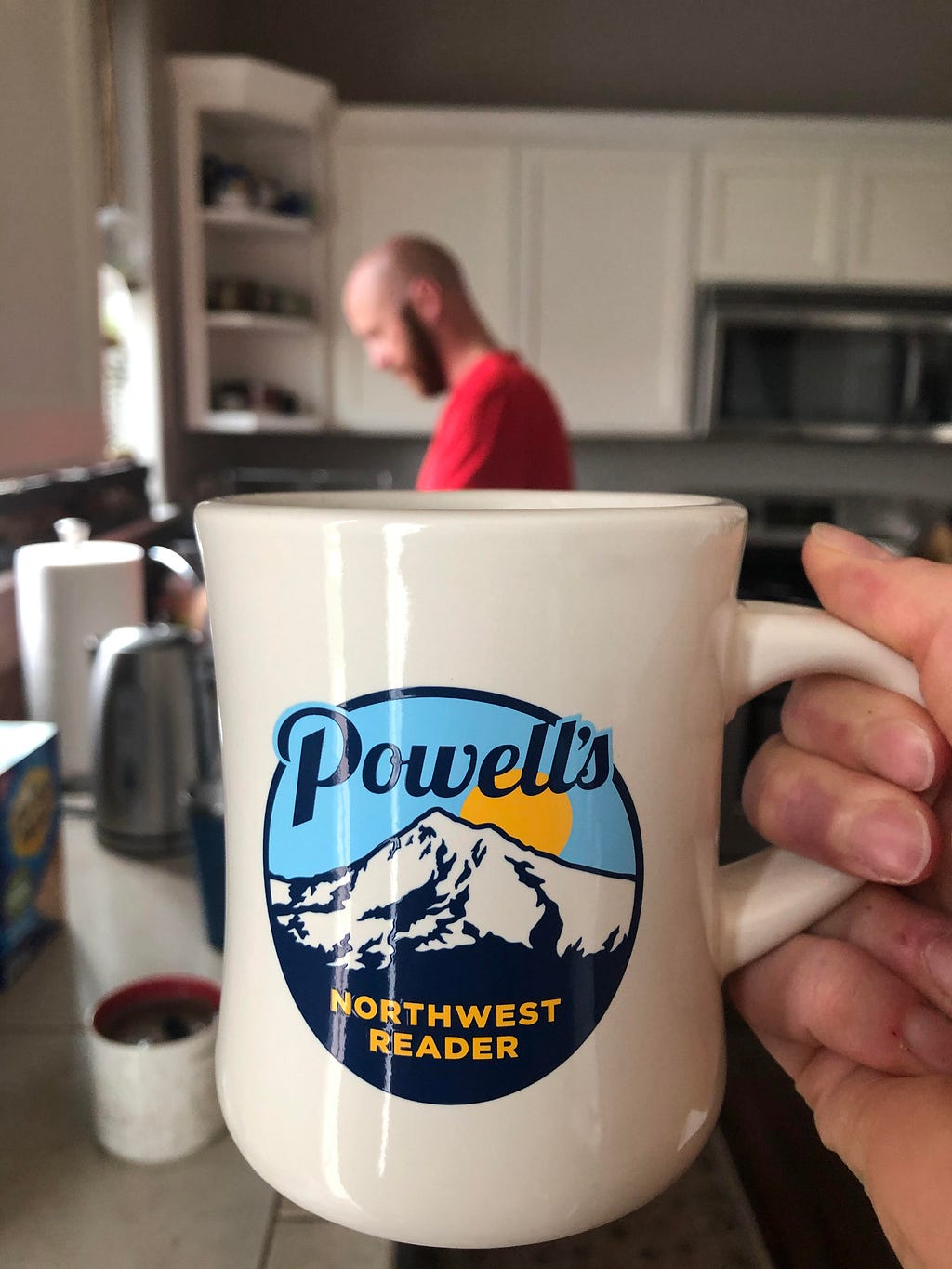 A hand holds a cream diner mug that says “Powell’s Northwest Reader.” Behind, a man in a red shirt stands at the sink