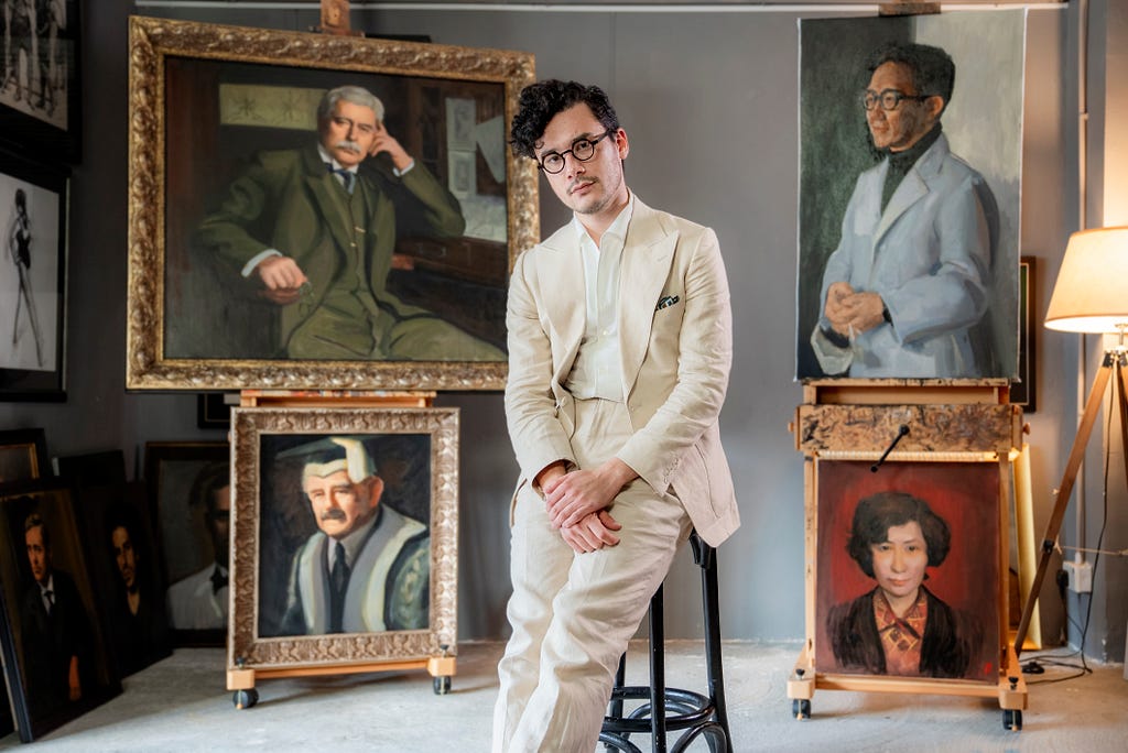 Artist Brendan Fitzpatrick sits on a stool in his studio in front of four portraits