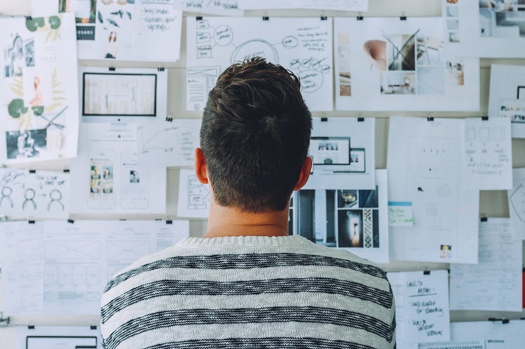 A person looking at a wall covered with paper sheets of paper containing sketches and annotated pictures.