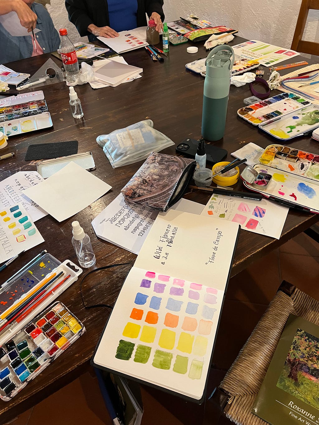 Photo of the worktable during a watercolor lesson by artist Roxanne Steed. Scratch papers show the gradients of different pigment concentrations in pinks, blues, oranges, and yellows.