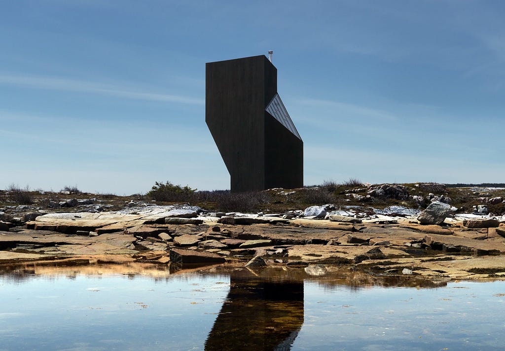An angular, black, wooden-clad building sits alongside a rocky coastline.