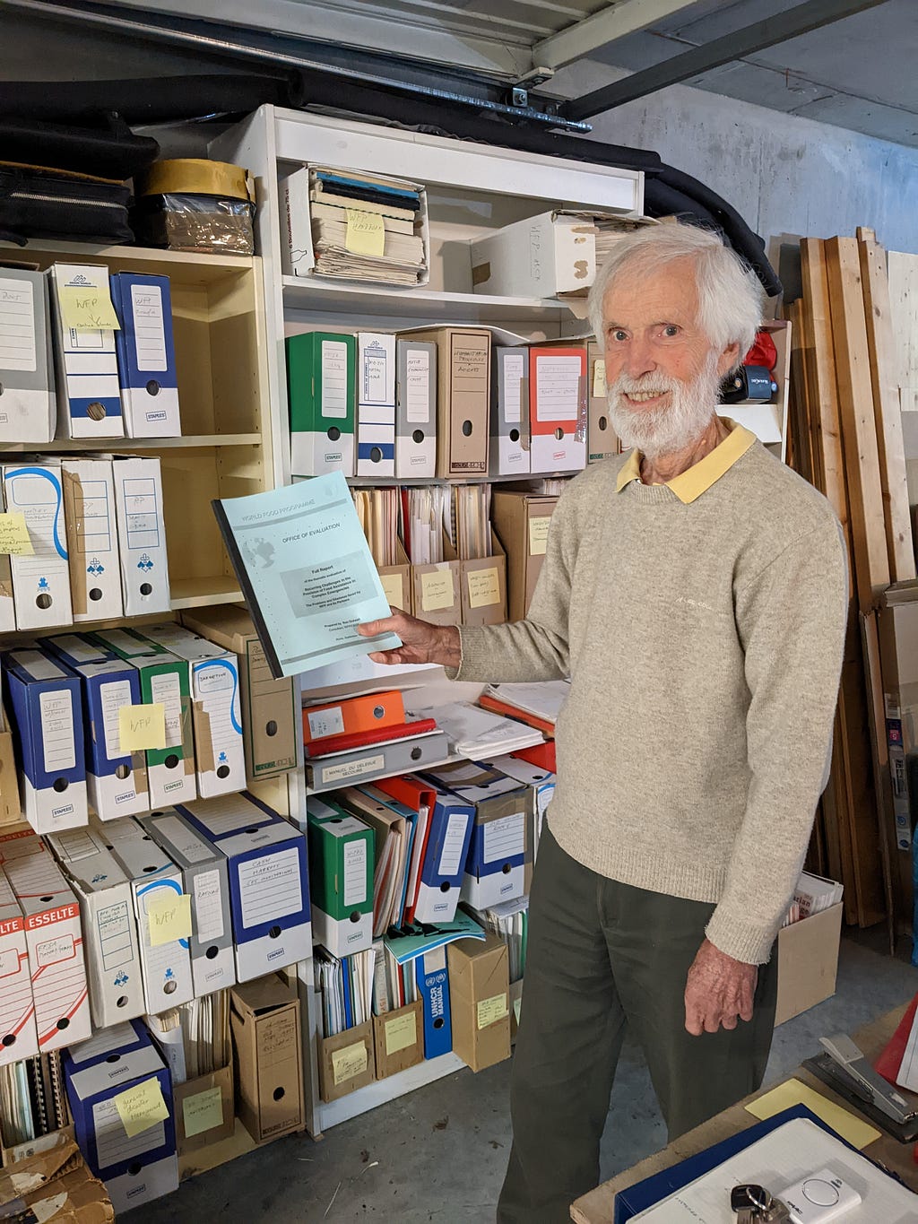 A photo of Ron Ockwell, looking at the camera and smiling. He is standing in front of two shelves, both full of boxes of documents. documents. In his right hand he is holding a report on “Recurring challenges in the provision of food assistance in complex emergencies” that he prepared for The World Food Programme in 1999.