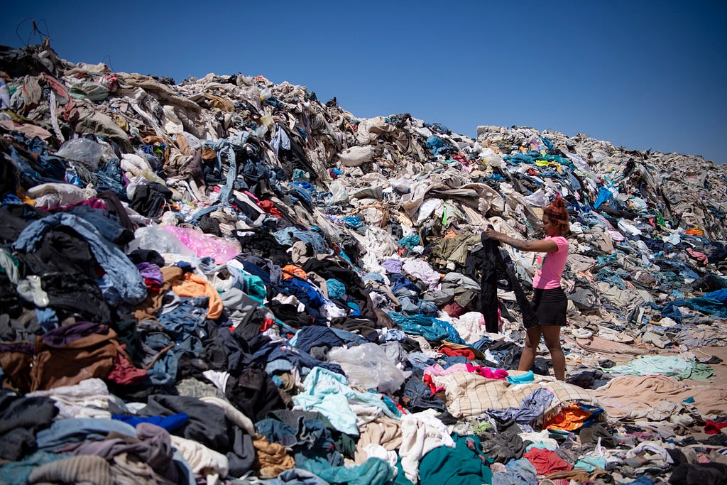 In the middle of a landfill full of clothes is a person holding up a garment as if they are interested in taking it home.