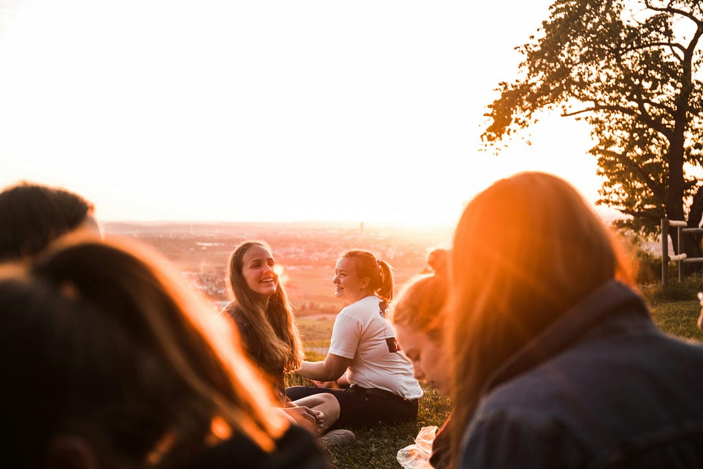 A group of young people sitting on the grass, with the glaring sunset behind them.