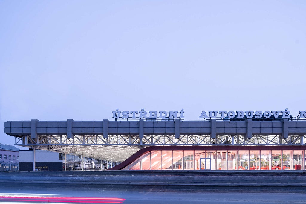 The exterior of the Zvonarka Bus Terminal. The the name of the station in Czech in large sign letters sits above the concrete roof. We can see the curve of the red cover swooping up from the ground to just below the concrete roof with the steel structure sitting between them.