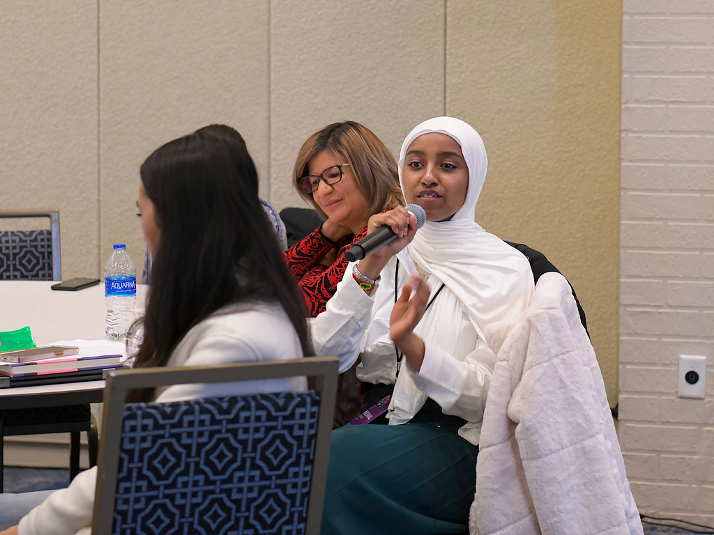 A group of young women sitting around a table. In a hijab, Newal N. of the Asian Pacific Development Center cohort holds a microphone to share her story during the New American Youth Leadership Council Summit at the National Immigrant Inclusion Conference 2022