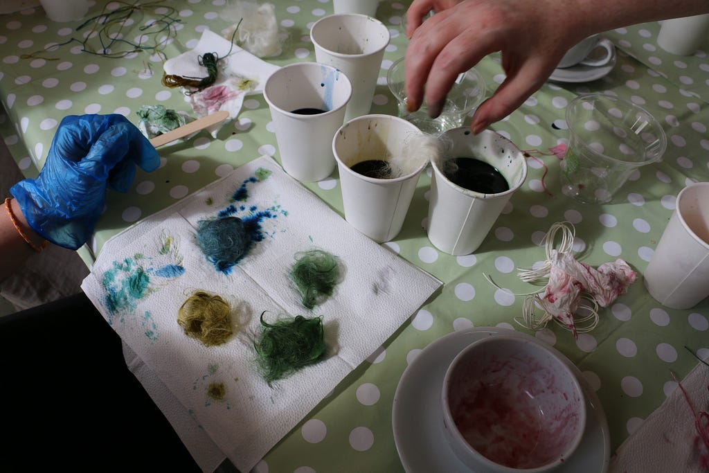 Clumps of yarn dyed blues and green sit on a paper towel with paper cups and dyes next to them.