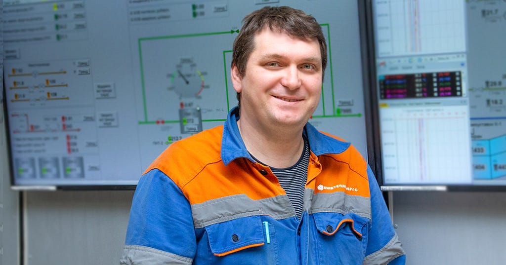 A man in an orange and blue engineering uniform stands in front of a technical drawing.