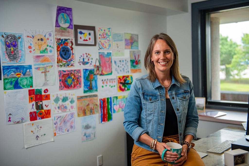 A 40-year-old woman sits on the edge of her desk holding a cup of coffee and smiling at the camera. A wall of children’s art is shown behind her.