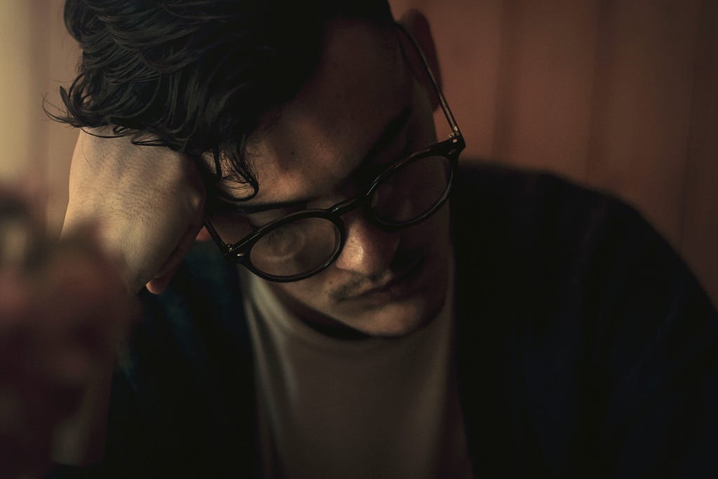 A man wearing glasses rests his head against his fist while looking downward.