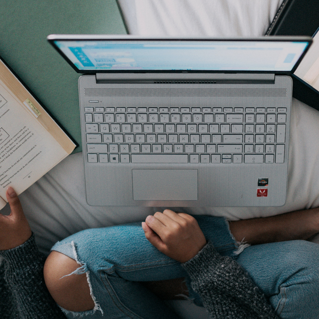 A woman, waering wripped jeans, sitting on a bed with her laptop open, a book to her right open and what seems to be a note pad and a folder on the bed as well. She appears to be researching something.