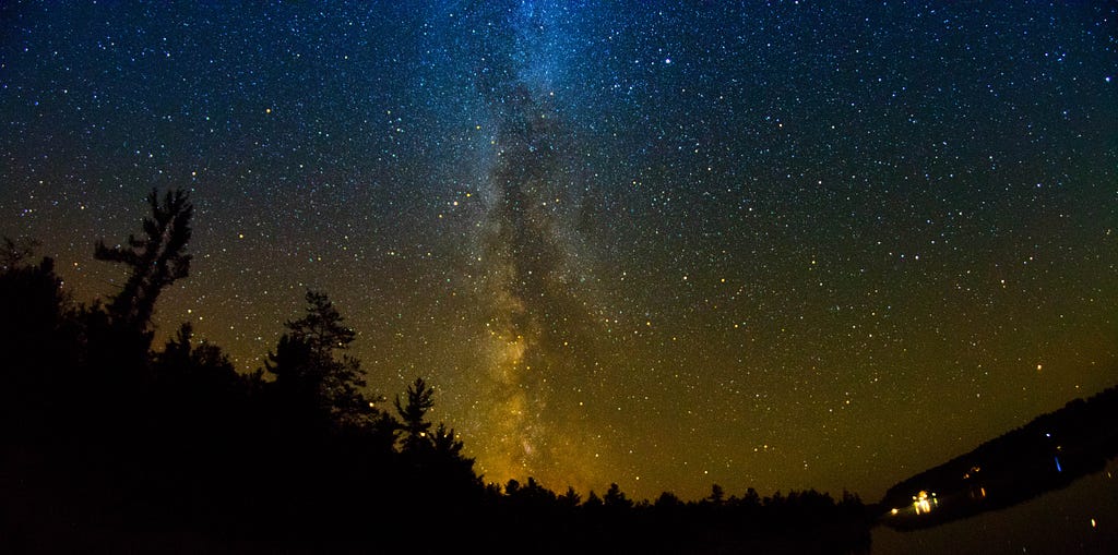 Photo of a night sky, trees line the bottom of the image, small lighted homes in the bottom left corner.