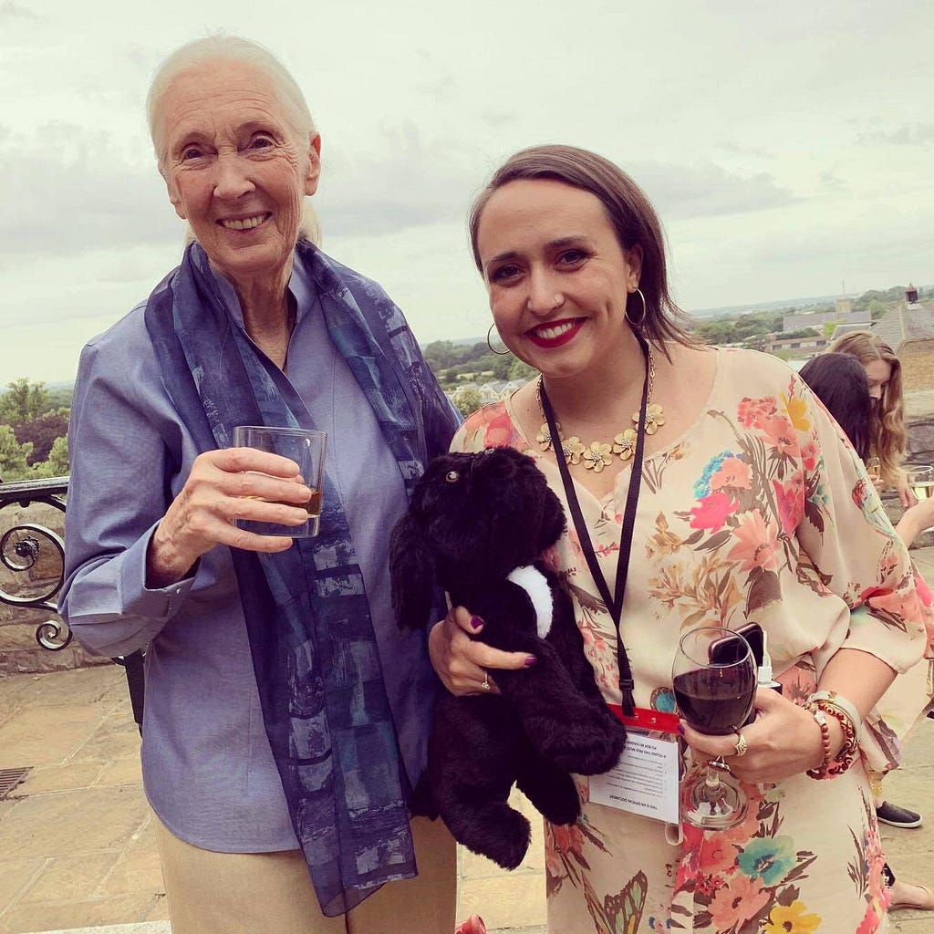 Jane Goodall stands in blue next to Emily Major in a creamy, floral shirt outside on an overcast day; both smiling wide for the camera.