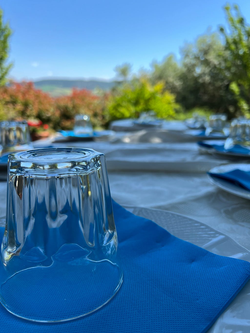 Close up photo of an outside lunch table with glasses, blue napkins, and while tablecolths. In the background are the far hills of the Val d’Orcia.