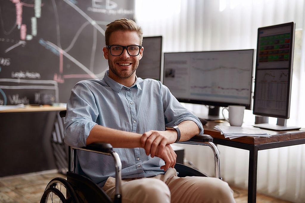 A young man with short, light brown hair and a beard is sitting in a wheelchair, smiling at the camera. He is wearing a light blue button-up shirt and beige pants. He is in a modern office setting with two computer monitors on a wooden desk behind him displaying charts and data. A chalkboard with various drawings and notes is visible in the background, along with a window covered by vertical blinds allowing natural light to illuminate the room. The man appears confident and relaxed in his profes