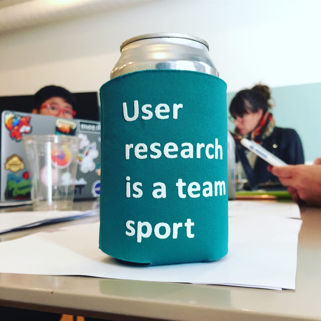 A soda can in a coozy that says “User research is a team sport,” sitting on a table with people & laptops in the background.