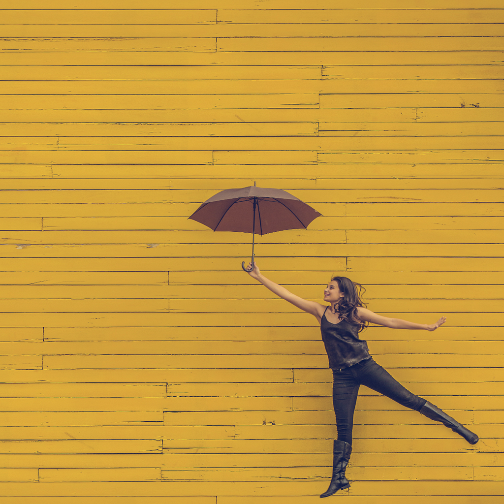 A woman, on the right of the photo, leaping with an umbrella in hand. All in front of a mustard yellow backround that resembes planks of wood.