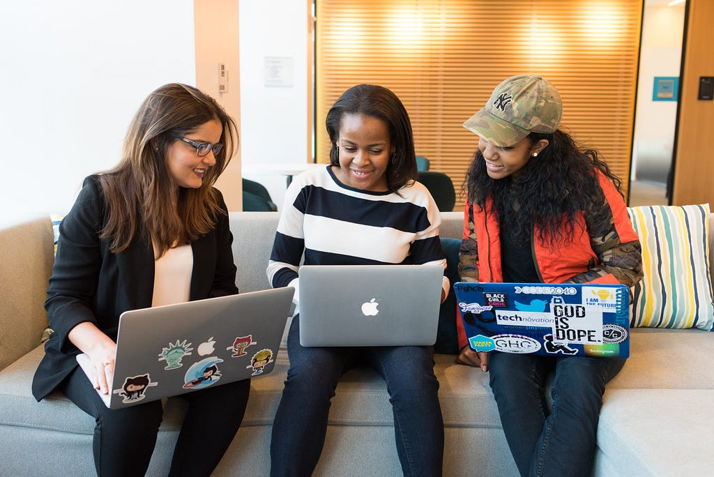 Three diverse women in technology sit on a couch with laptops,  working together on technology projects, mentoring each other