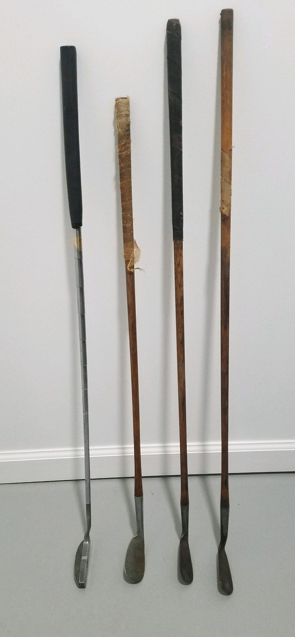 Picture of four golf clubs. From left to right, a putter from the 1970’s or 1980’s, and three old clubs from Scotland. They have wood shafts, very worn grips, and forged iron heads. From the Ross family archives, photo by author.