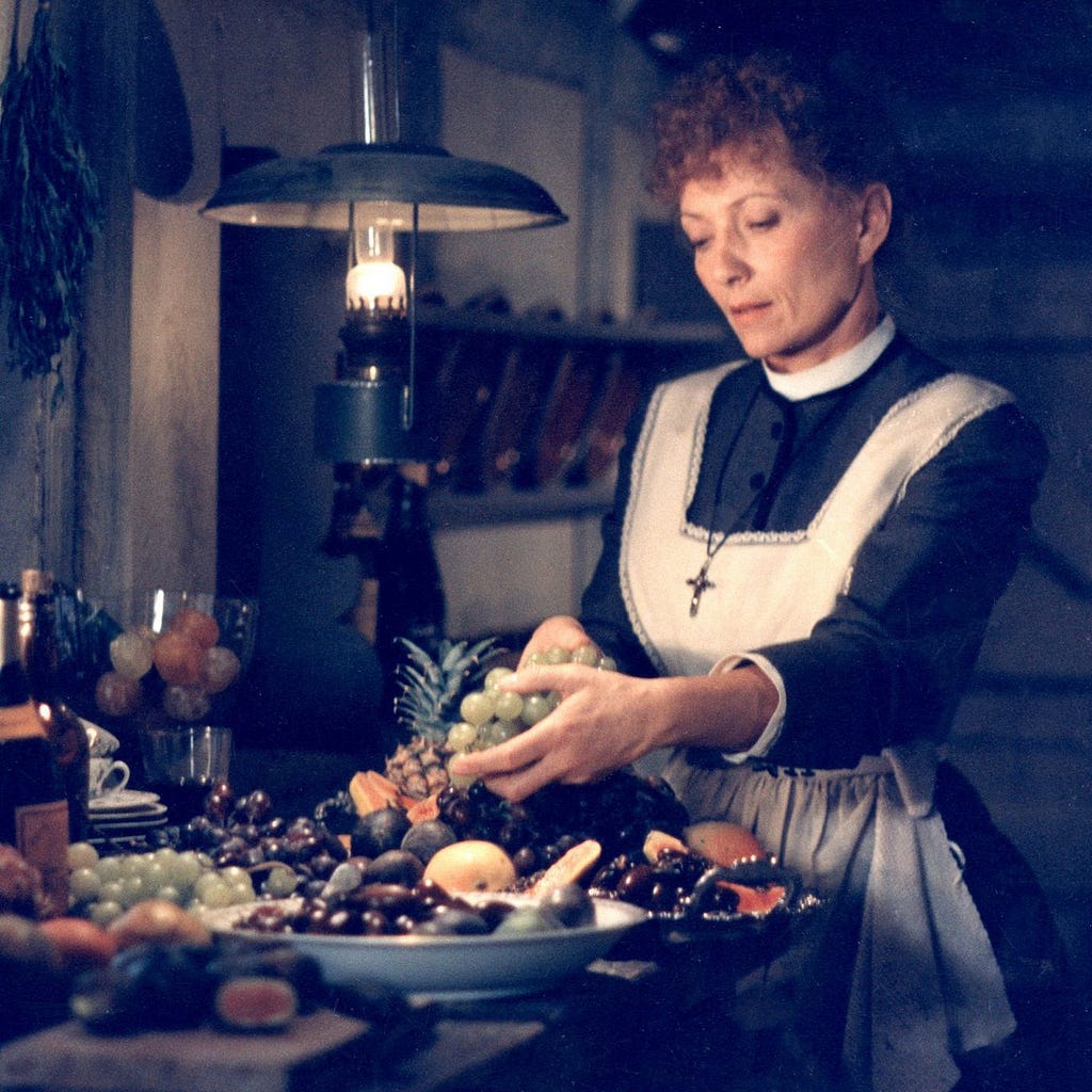 An austerely dressed woman arrange a lavish platter of fruit in a dim, old-fashioned kitchen.