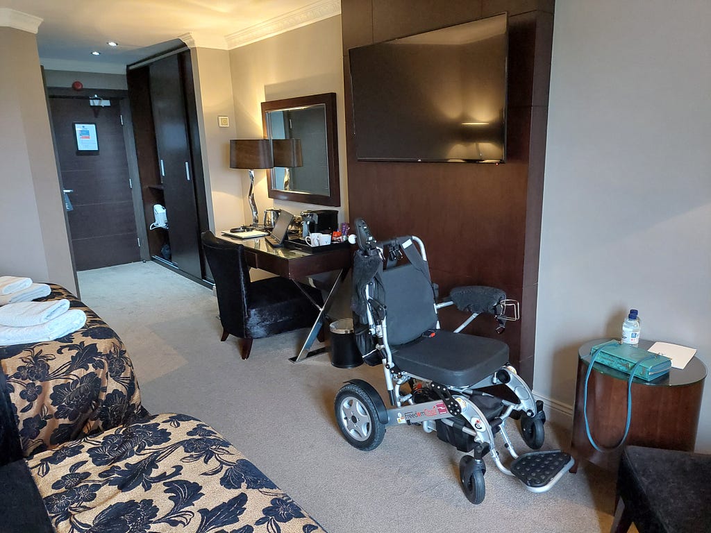 The hotel room shown from the windows, looking towards the door to the corridor. From left to right, there is a wardrobe with sliding doors, dressing table with low chair, Claire’s electric wheelchair is under the flat-screen TV and there is a coffee table and second chair.