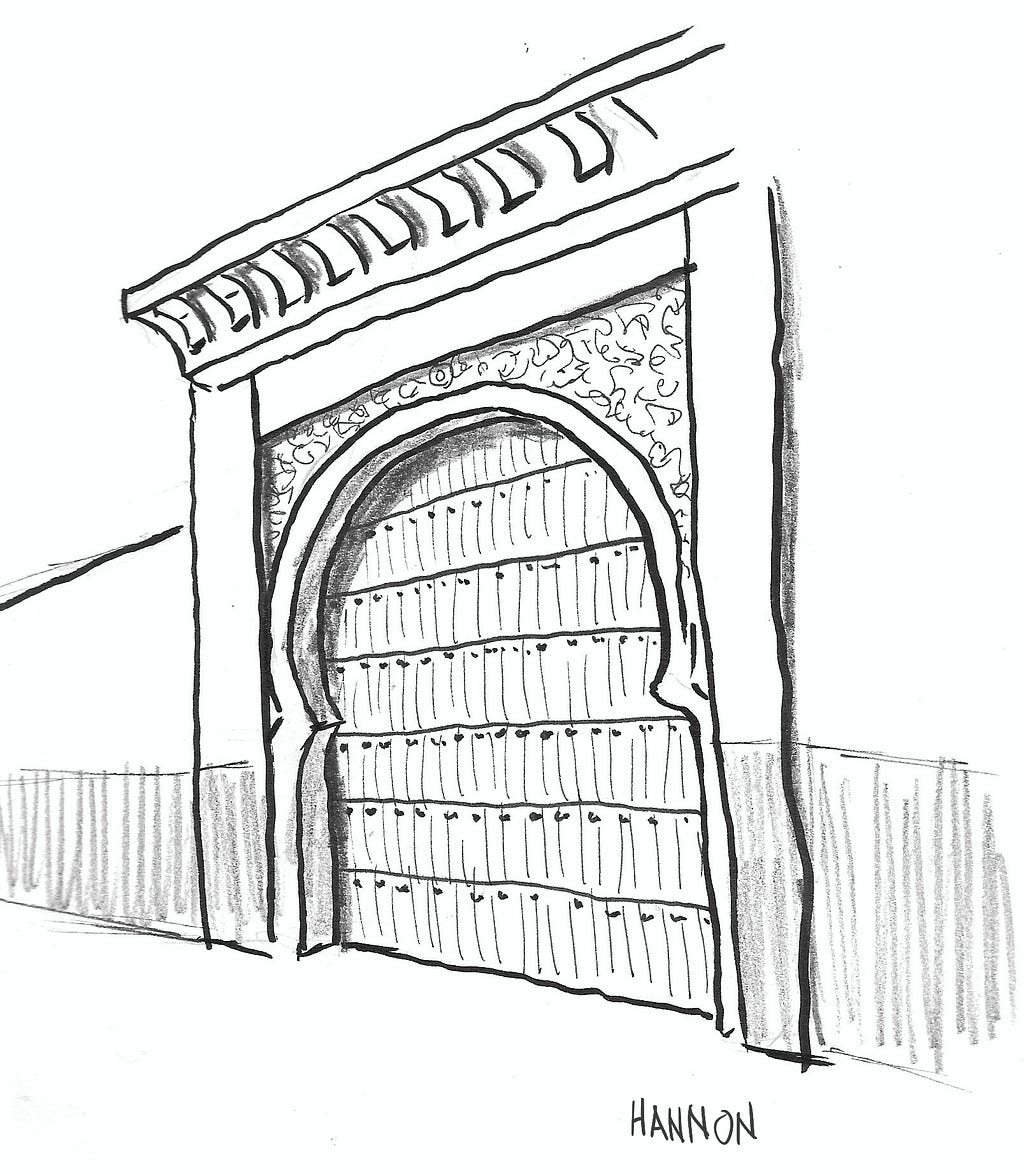 A doorway framed by heavy beams on the sides and top. Above the top beam is a large molding with a concave face and notches carved into it at regular intervals. Inside the outer molding is a smaller frame that is straight and vertical halfway up. The frame curves inward briefly, then opens up into a semi-circle. The door itself is made from several horizontal sections which are segmented into vertical slats.