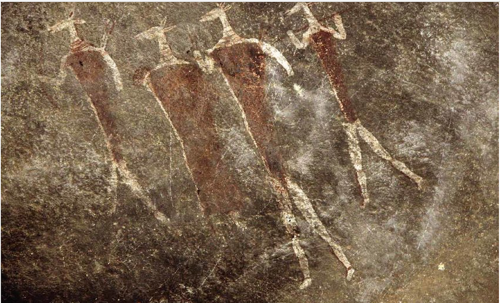 Stick figures in white of 4 creatures with heads like antelopes and body and feet like human, painted on brown rock surface.