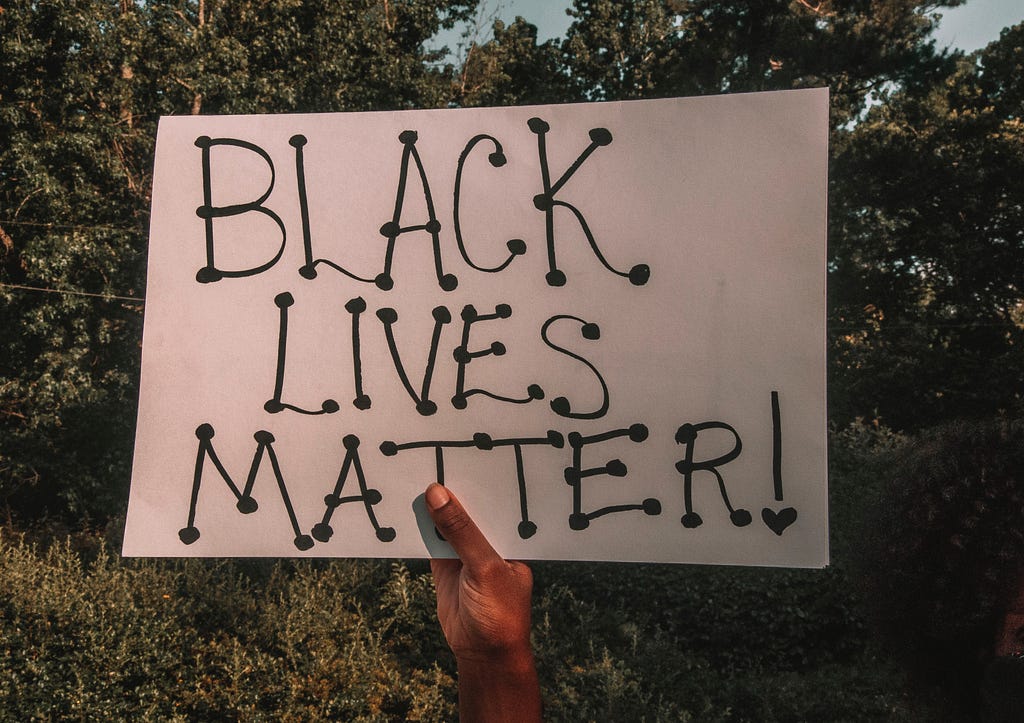 A black hand holds up a sign that reads “Black Lives Matter!”