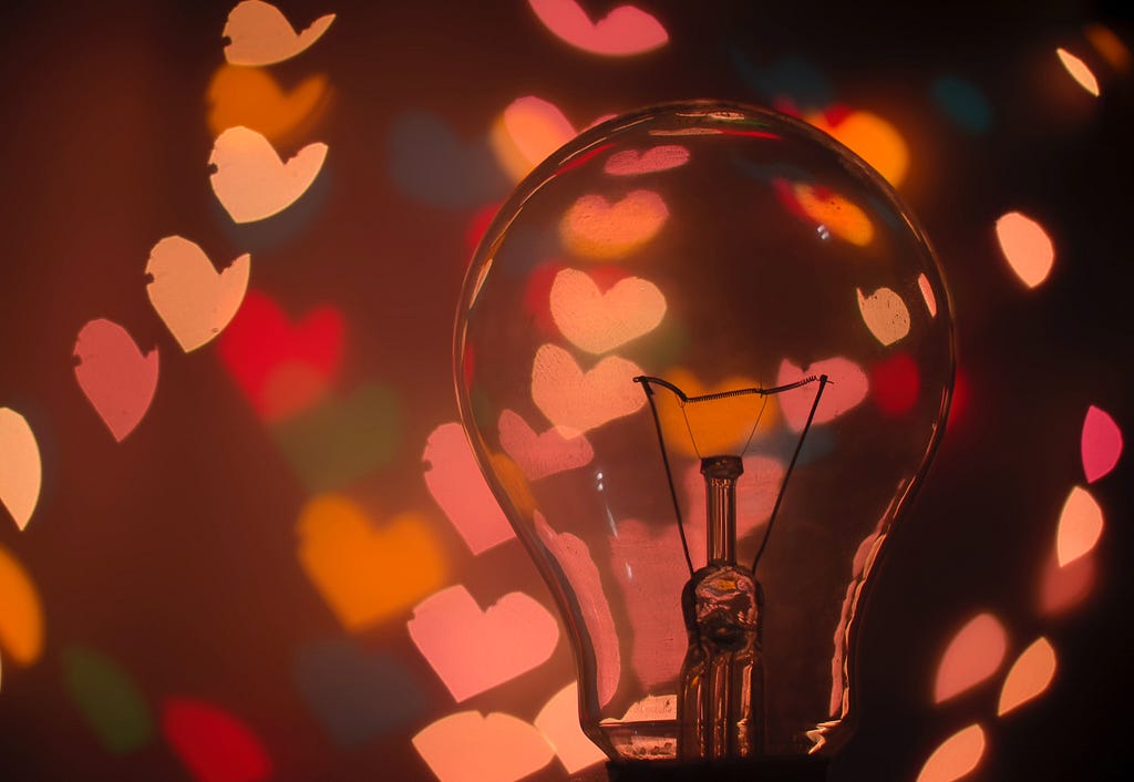 Close-up of a lightbulb with pale red lights in heart shapes surrounding it.