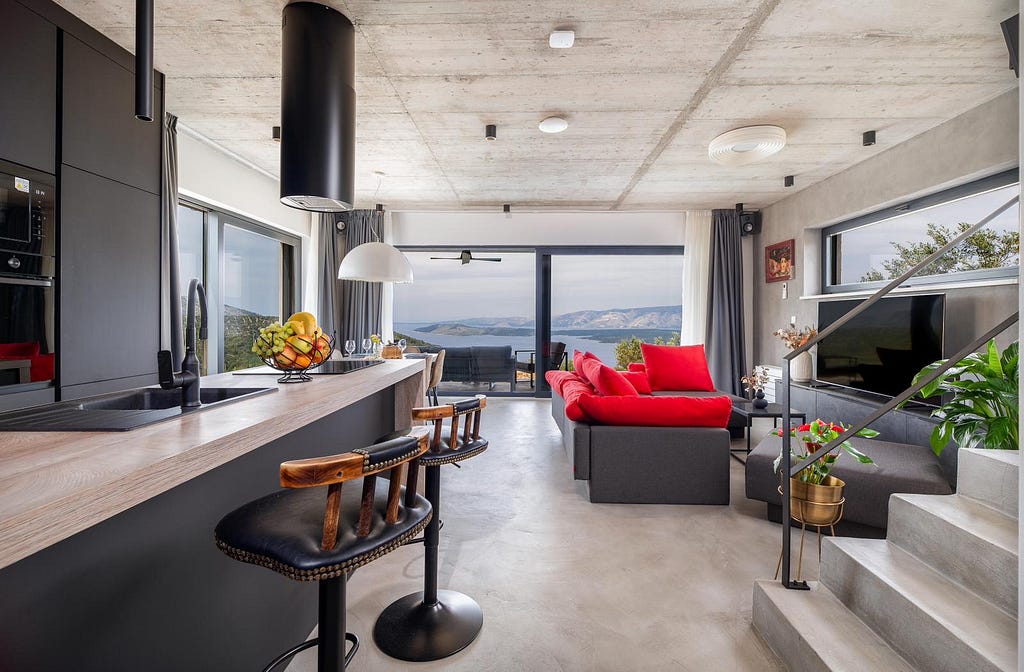 A contemporary open-plan living area with large windows offering stunning views of the sea and distant hills. The space features a sleek kitchen with a wooden countertop, black sink, and modern bar stools. Adjacent to the kitchen is a cozy living area with a dark gray sectional sofa adorned with vibrant red cushions. The room is tastefully decorated with indoor plants, a flat-screen TV, and stylish lighting fixtures.
