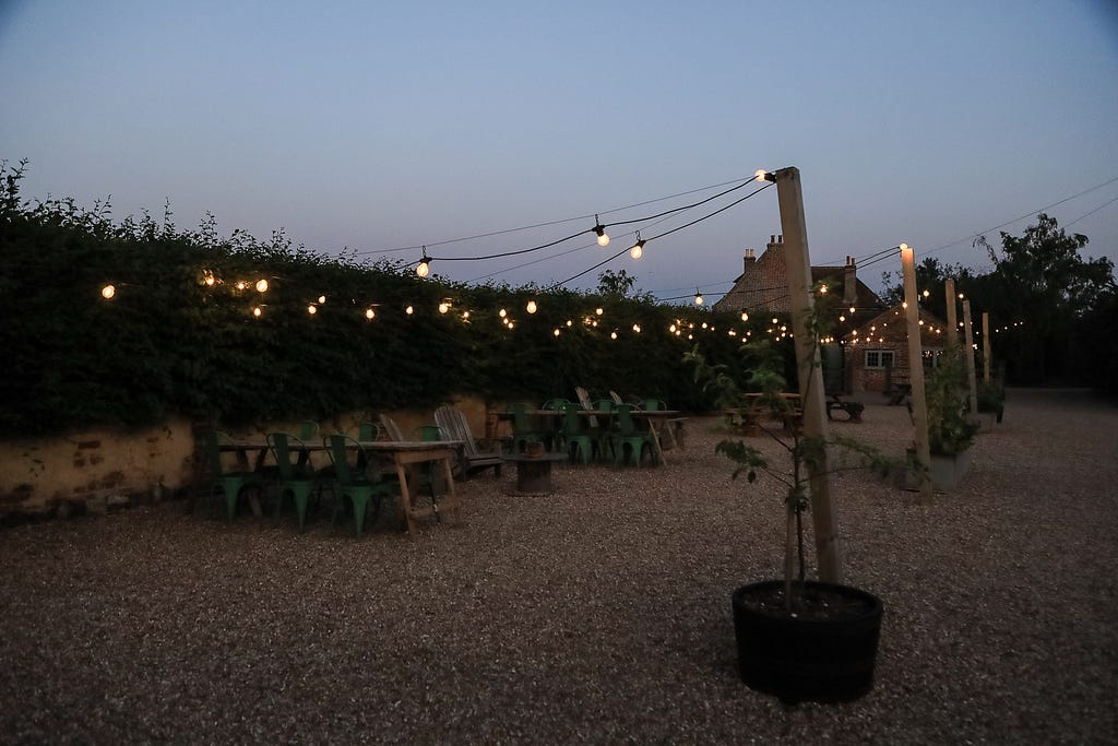 A dark picture of the front of the elmley grounds at night. There are tables and chairs set out with no one on them. The floor is gravel. There are strings of fairy lights over head.