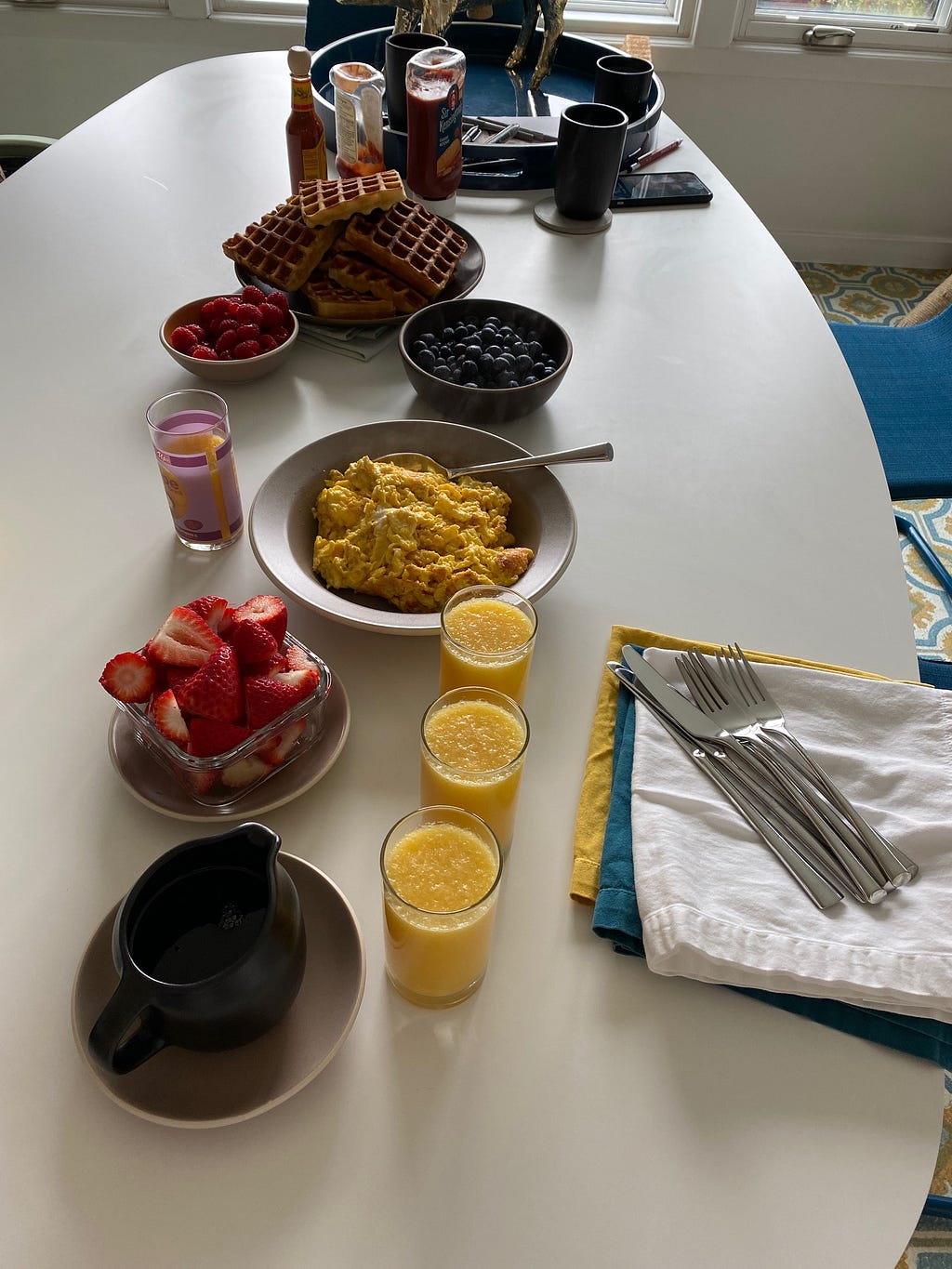 A sumptuous “insourced” brunch in a home dining area with fresh berries, scrambled eggs, homemade waffles and glasses of orange juice on a white table.