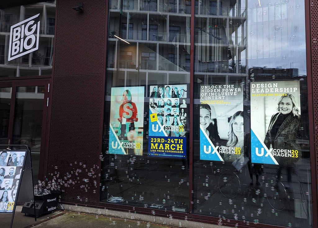 Digital movie posters outside the conference venue called Big Bio, a new cinema building. The poster shows a black and white image of me and Demi smiling, with the title of our workshop ‘Unlock the hidden power of inclusive language’, and the UX Copenhagen blue, white and yellow branding. There is a bubble machine next to the posters blowing bubbles across the posters (which were there to represent the conference theme of invisibility).