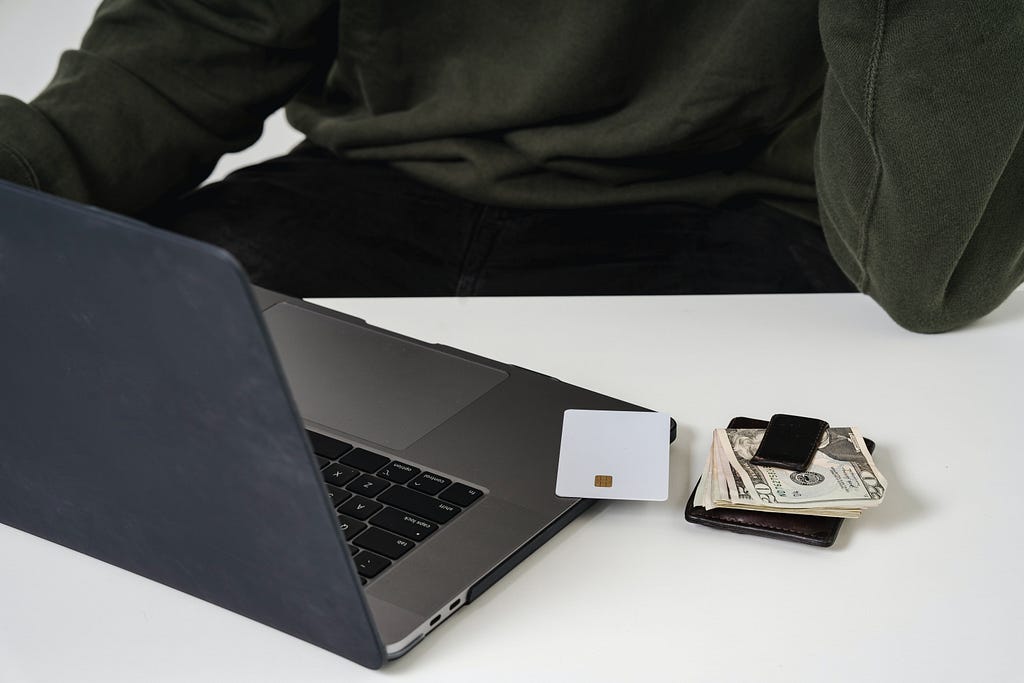 A person sits at a white desk, their wallet and credit card is laid next to his laptop. They are assumed to be shopping online.